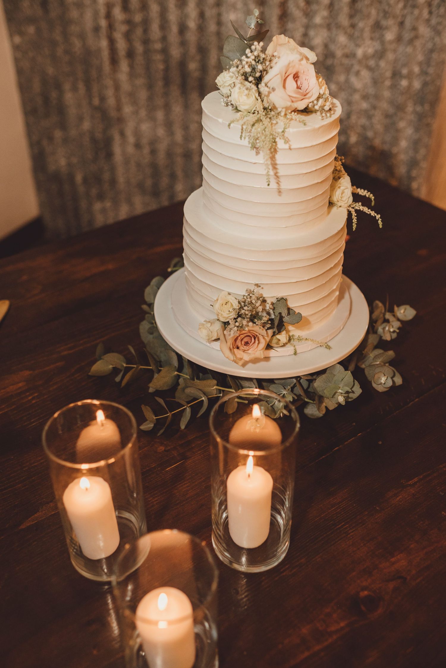 Elegant two-tier white wedding cake with blush roses, surrounded by glass pillar candles on a rustic wooden table.