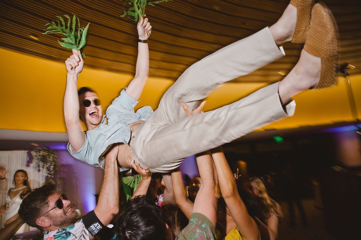 Man crowd surfing in white suit holding flowers at a lively wedding reception party.