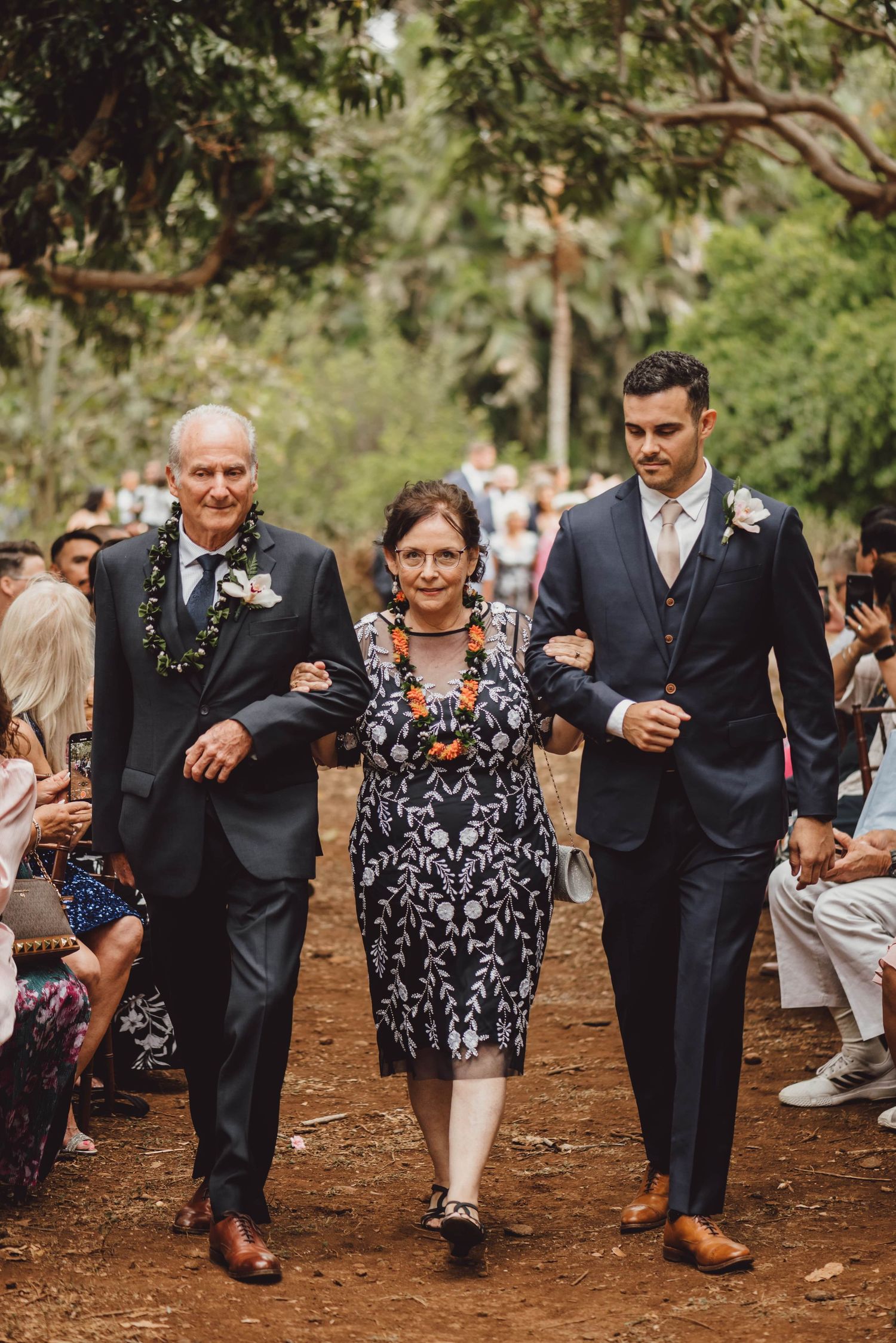 Wedding guests walk in a playful line through a wooded outdoor venue.