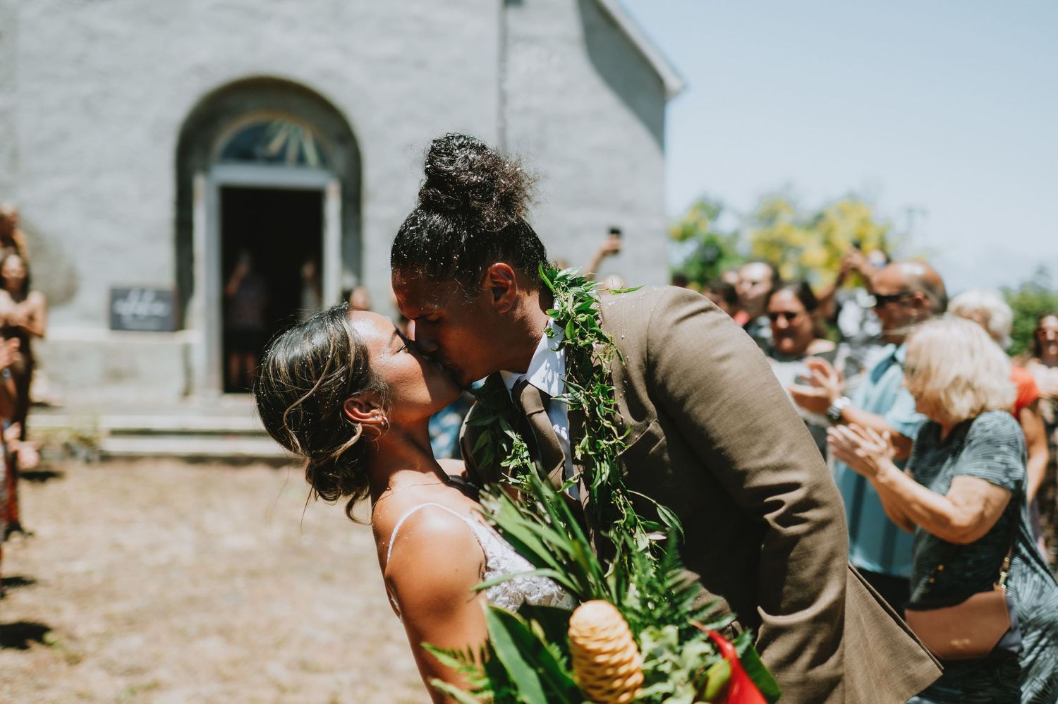 Wedding guests celebrate joyfully during an outdoor ceremony with tropical greenery and leis.