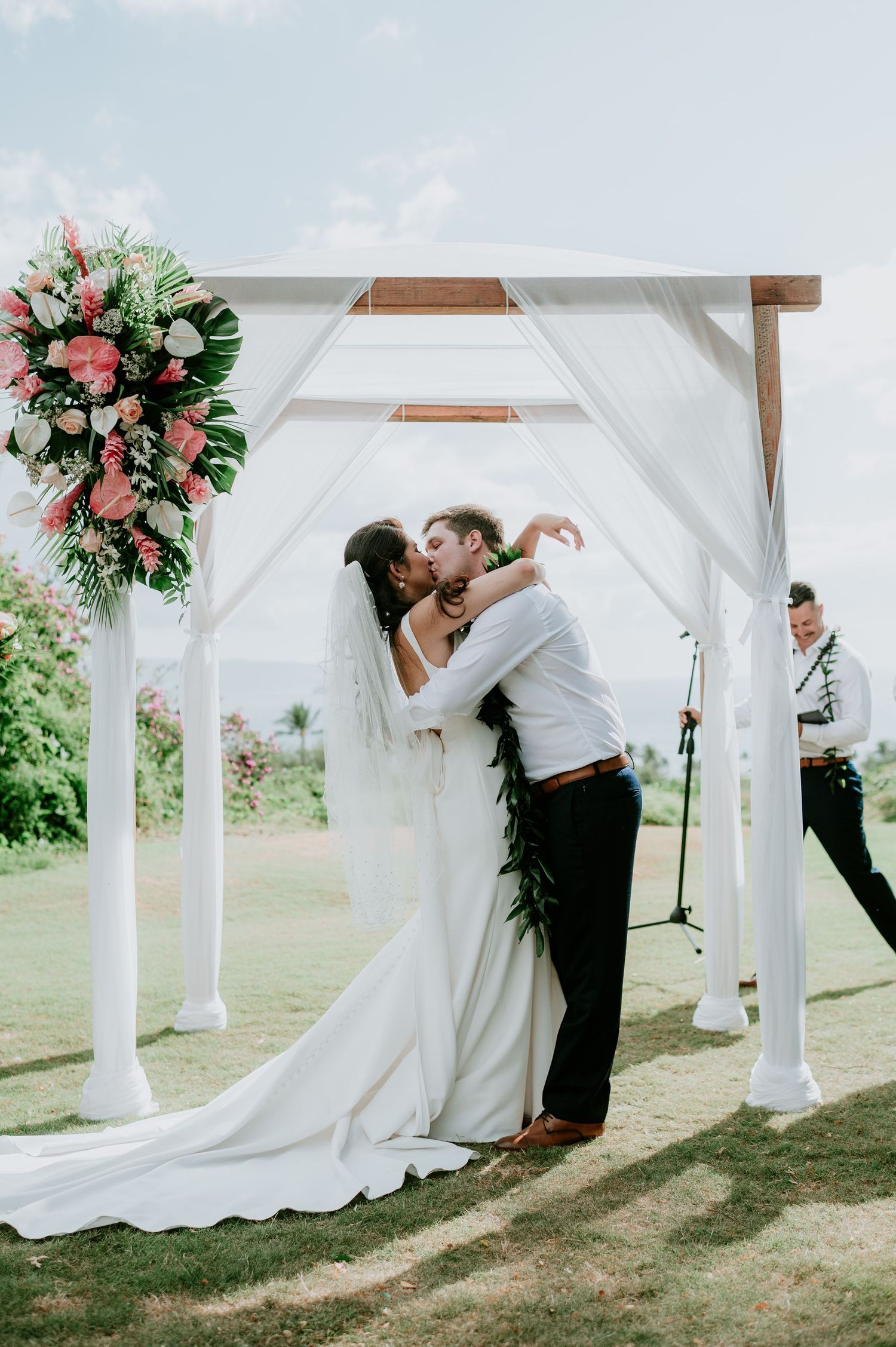 Newlyweds share their first kiss under an elegant white gazebo decorated with pink flowers.
