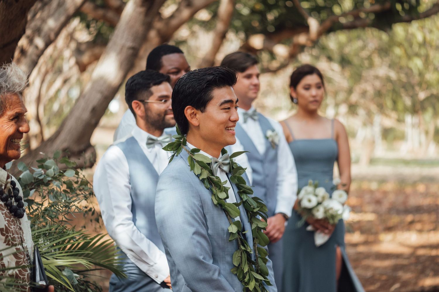 Groom wearing floral lei at outdoor wedding ceremony with wedding party in blue-gray attire under trees.
