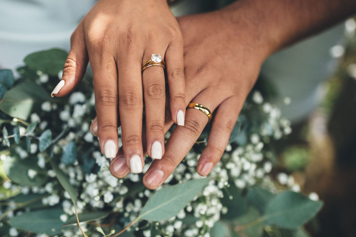 Couple displaying wedding rings on hands resting over eucalyptus and baby's breath bouquet.