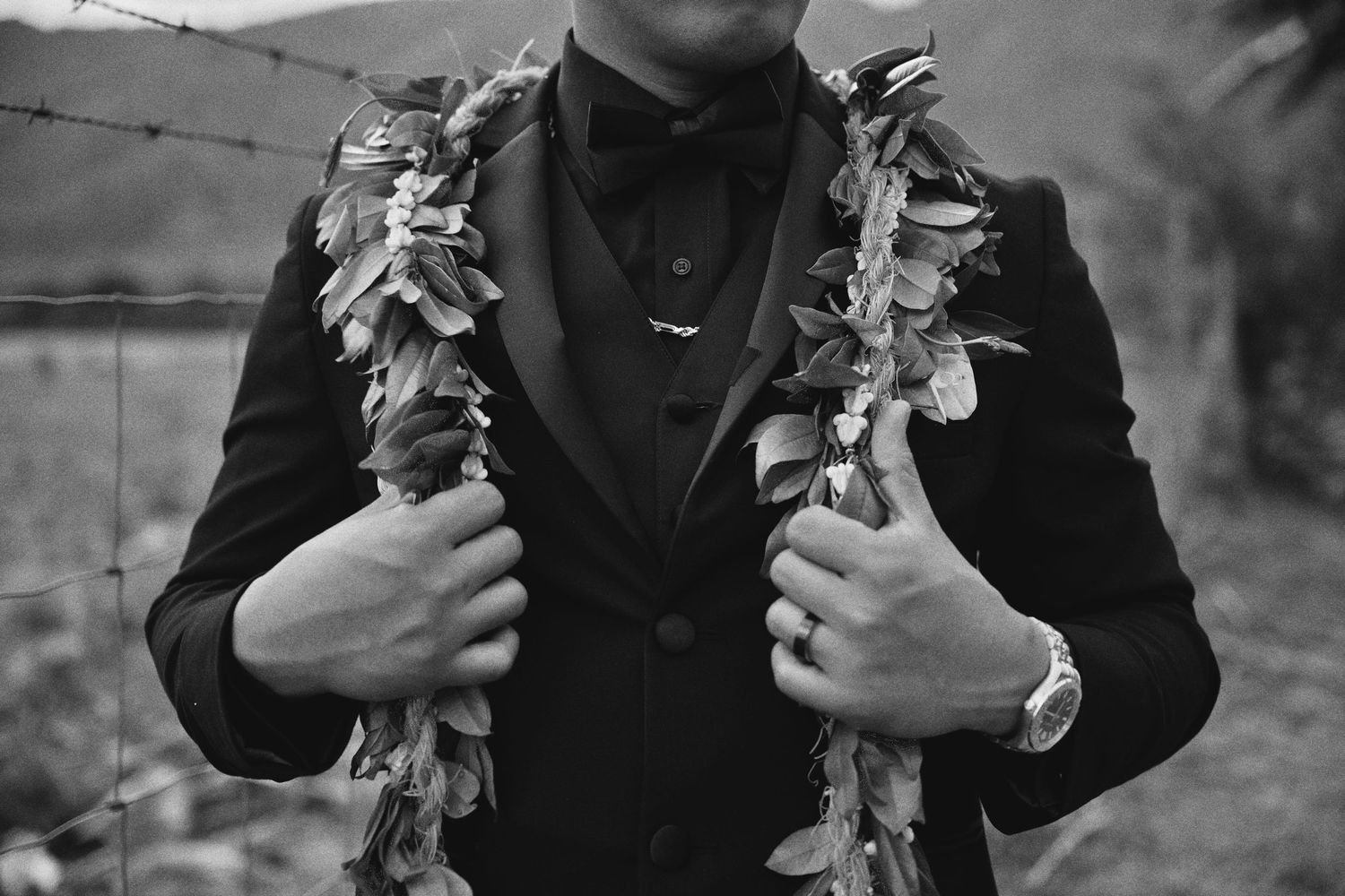 Black and white photo of a groom in a tuxedo wearing a floral lei, holding his lapels outdoors.
