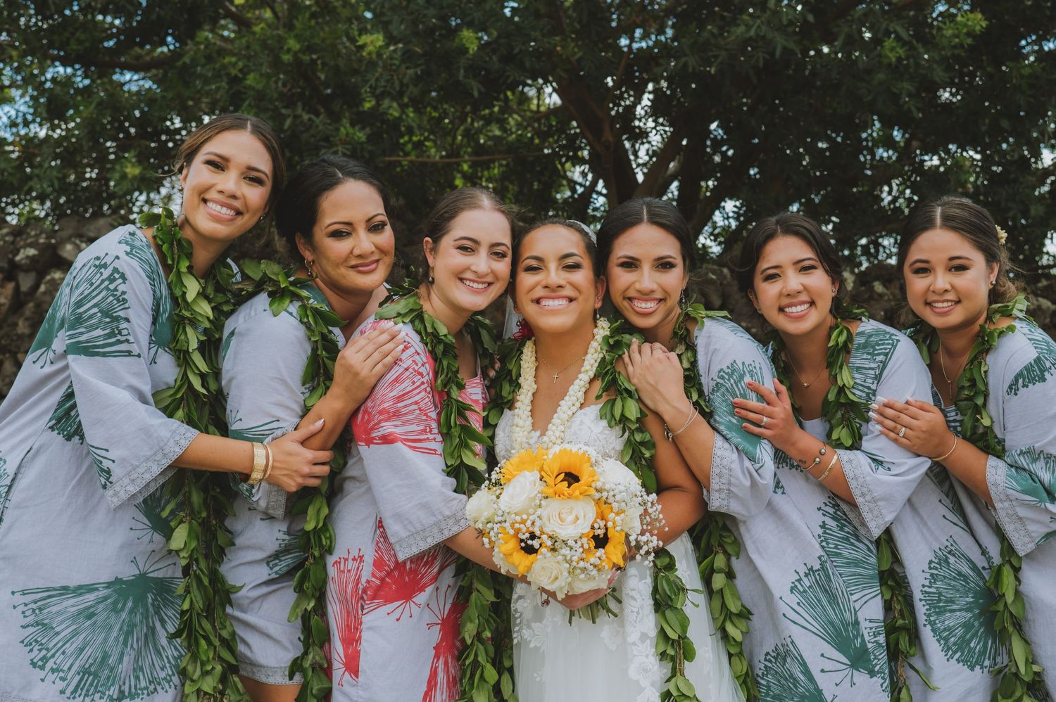 Bride holding sunflower bouquet poses with six bridesmaids wearing floral robes and green lei garlands outdoors.