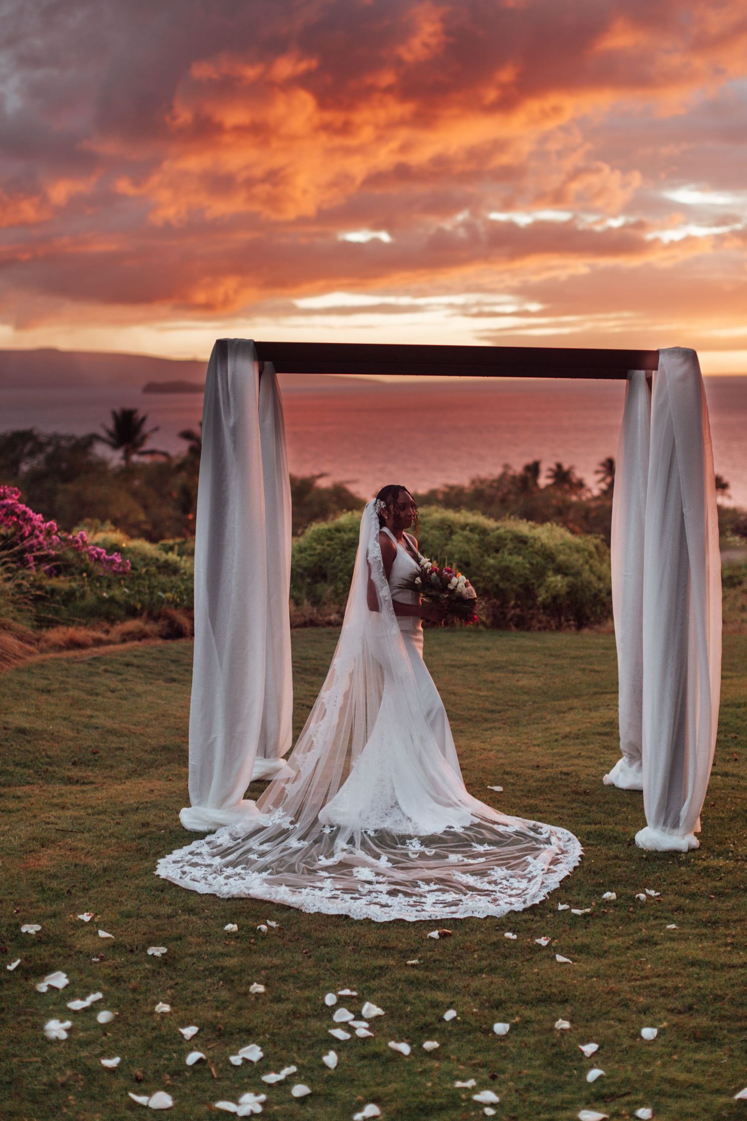 Bride in lace veil stands under draped wedding arch at stunning Hawaiian sunset ceremony overlooking the ocean.