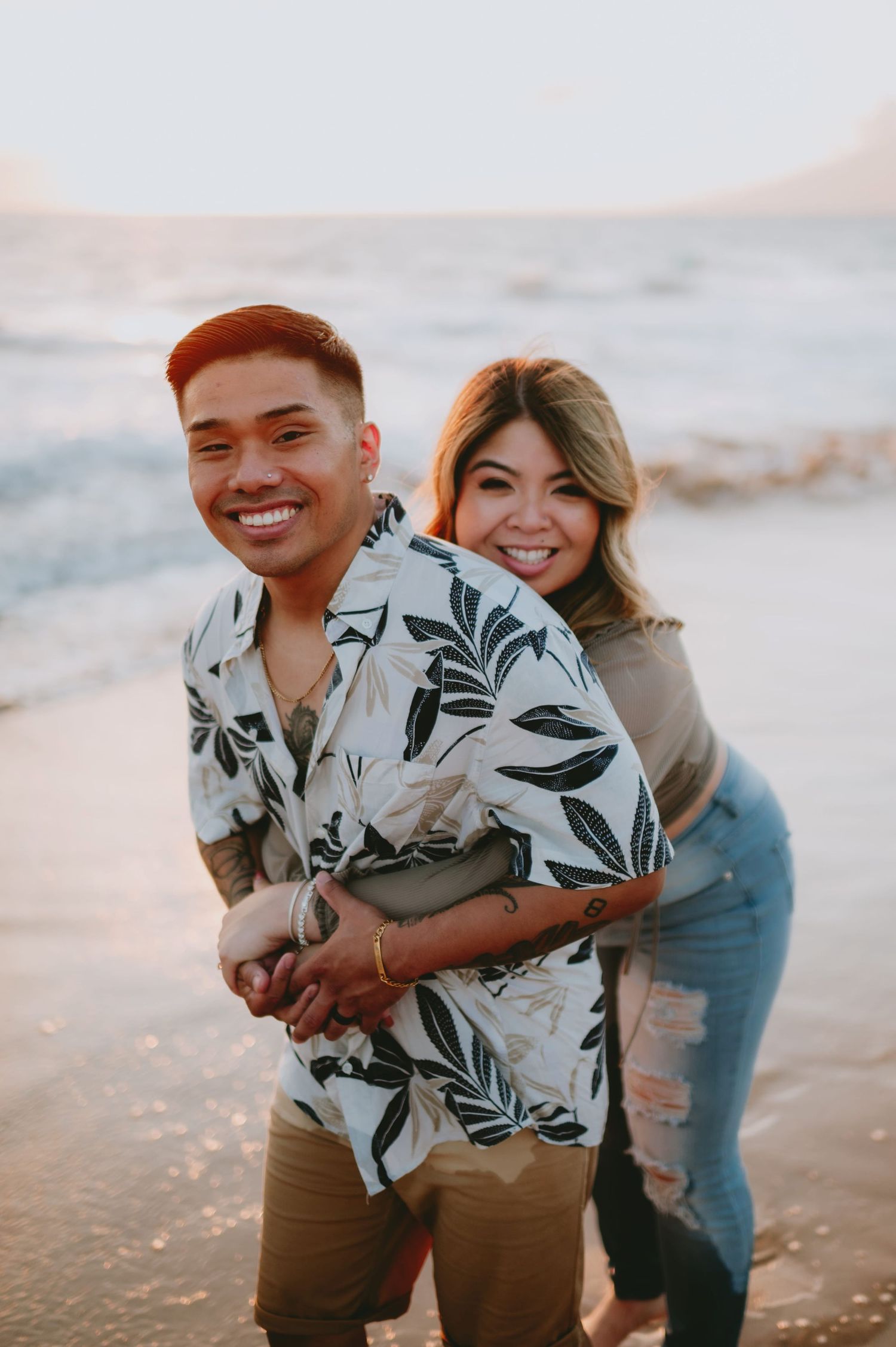 Smiling couple posing on beach at sunset with man in floral Hawaiian shirt.