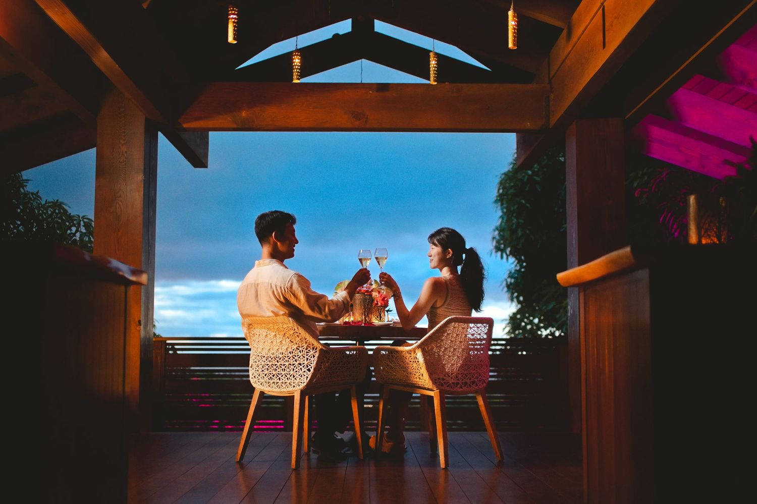 Couple toasting wine glasses at a romantic outdoor dinner table under a wooden pergola at dusk with ocean views.