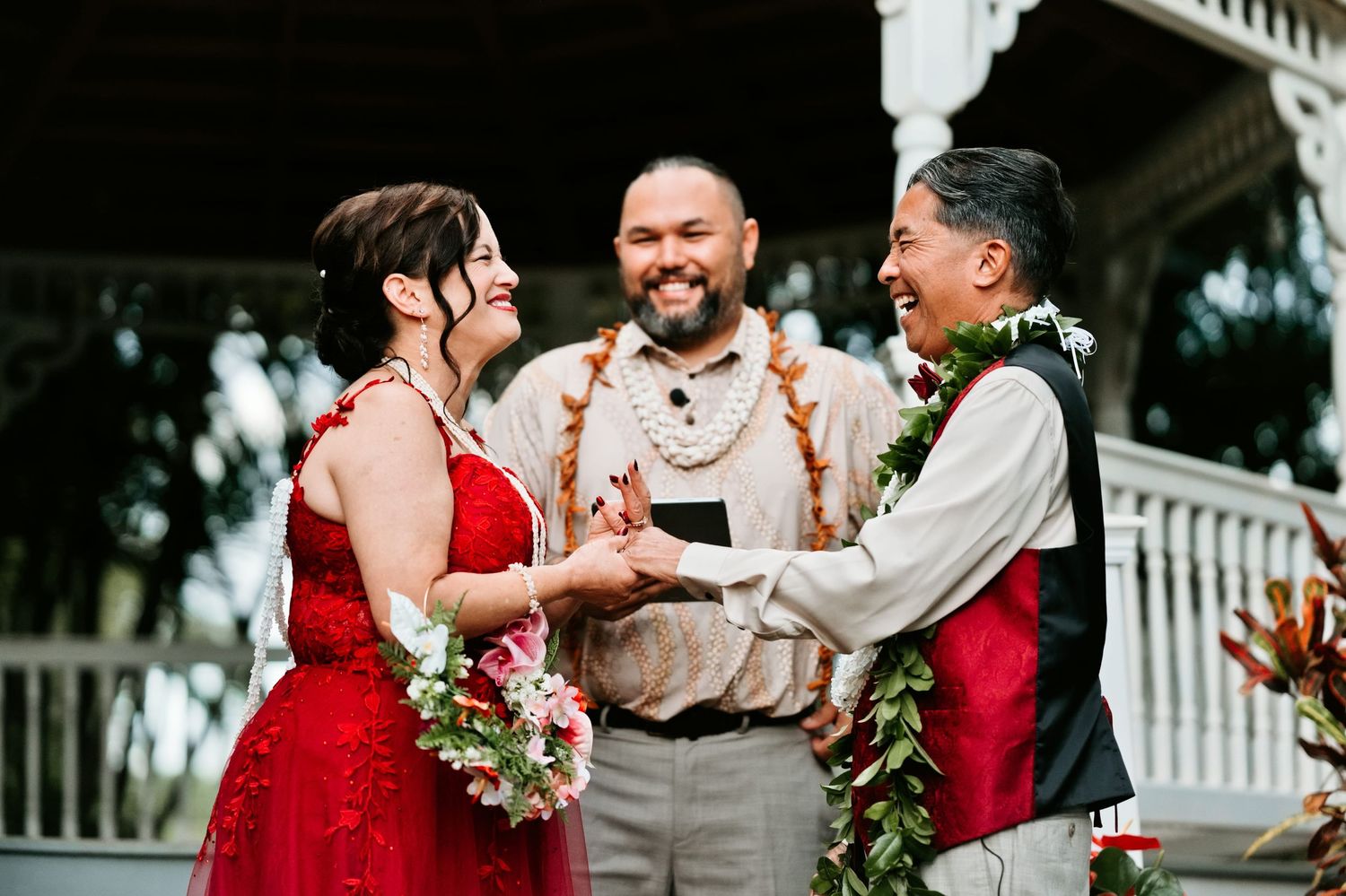 Couple exchanges vows at outdoor Hawaiian wedding ceremony with officiant, bride in red dress, groom wearing lei.