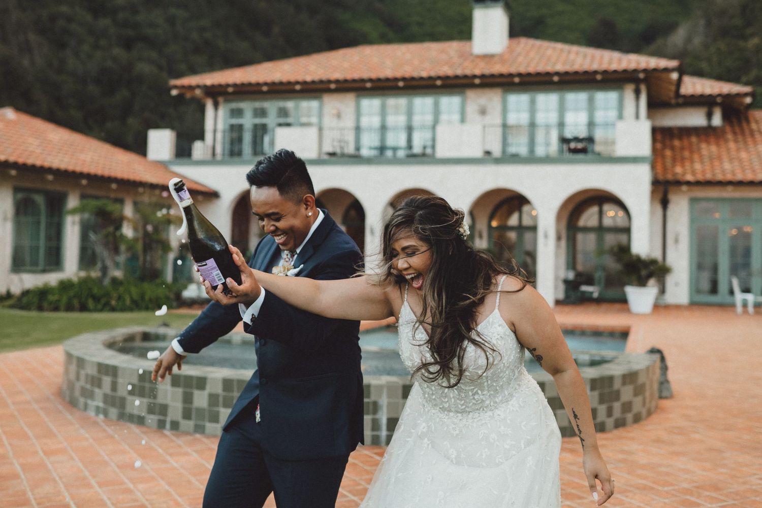 Joyful newlyweds celebrating with champagne outside a luxury villa, bride in white gown and groom in navy suit.