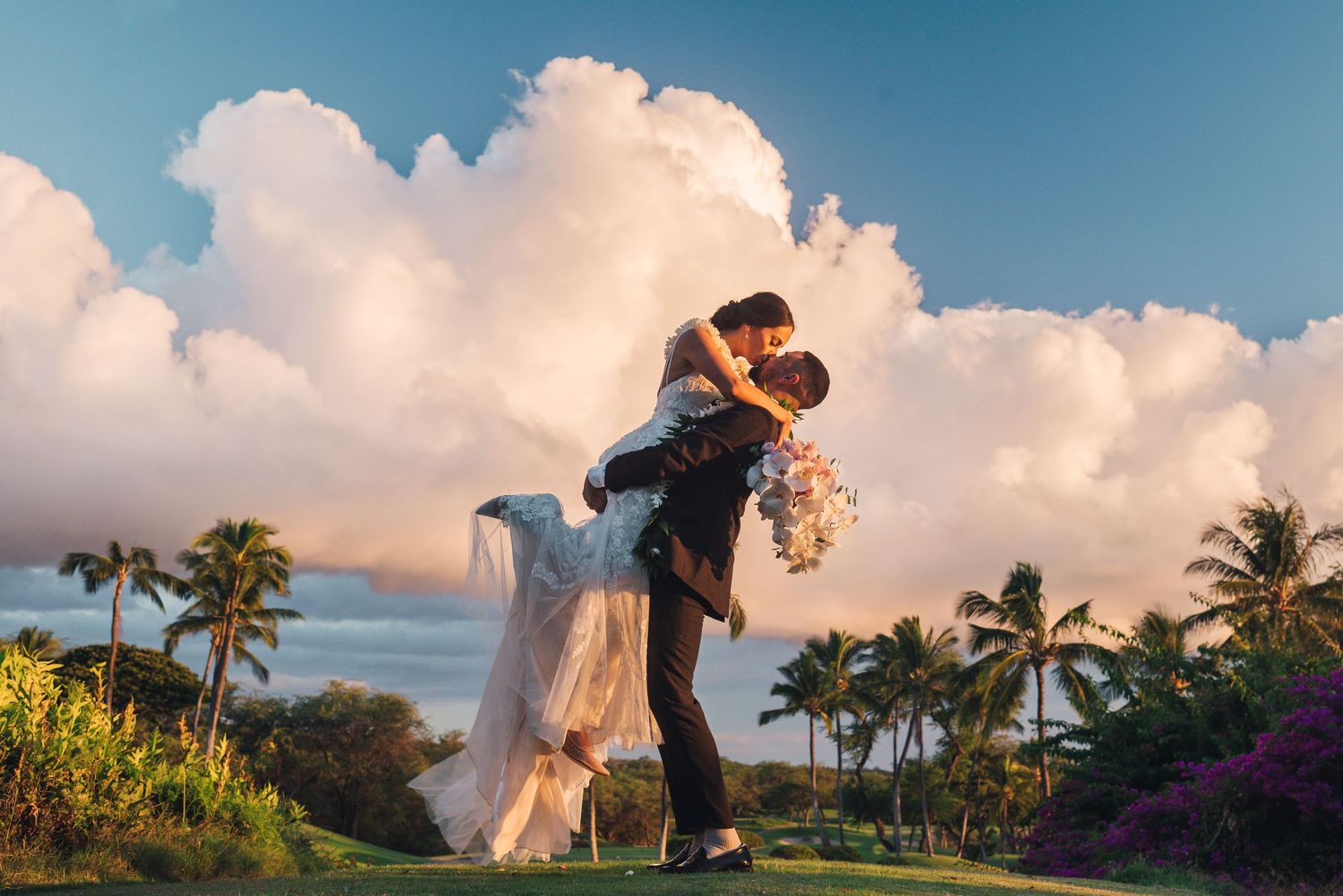 Groom lifts bride in wedding attire with tropical palm trees and dramatic clouds at a Hawaii wedding.
