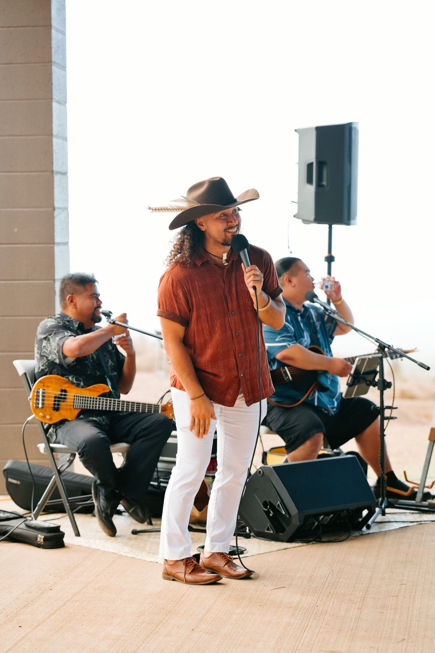 Live band performance with cowboy hat vocalist singing on stage with guitarist and drummer at outdoor event.