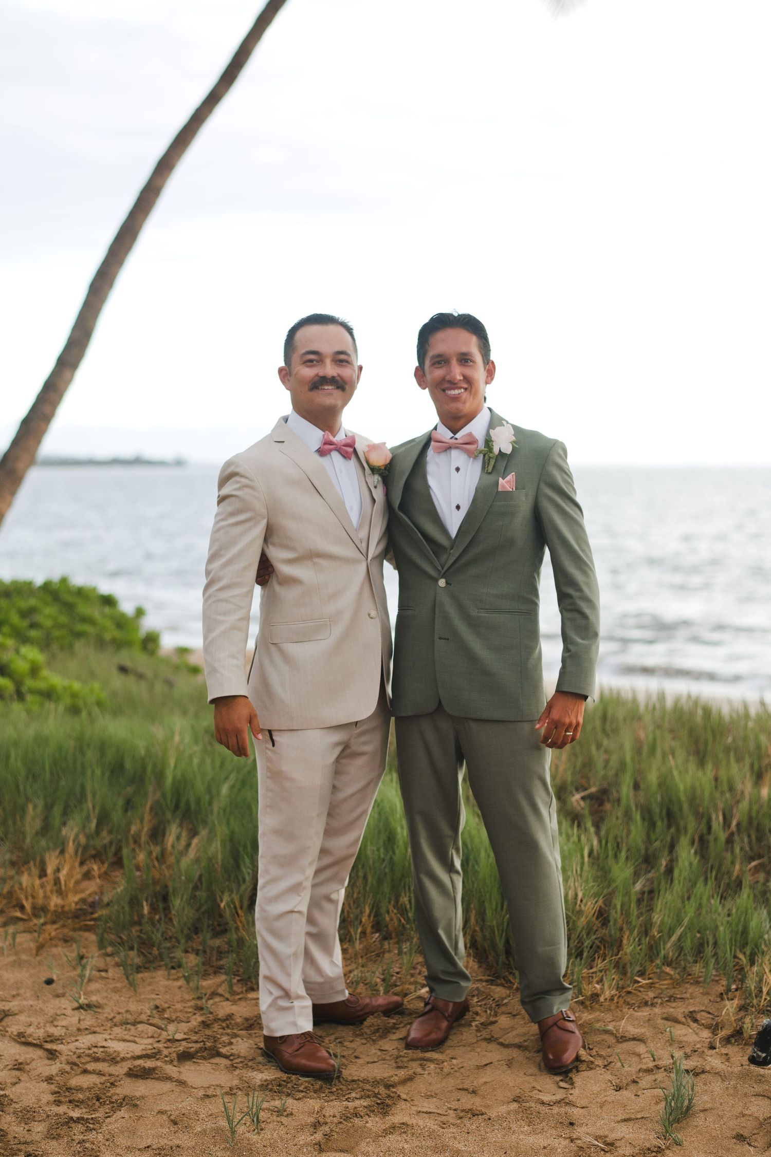 Two grooms in tan and green suits with pink bow ties pose on a tropical beach at their wedding.