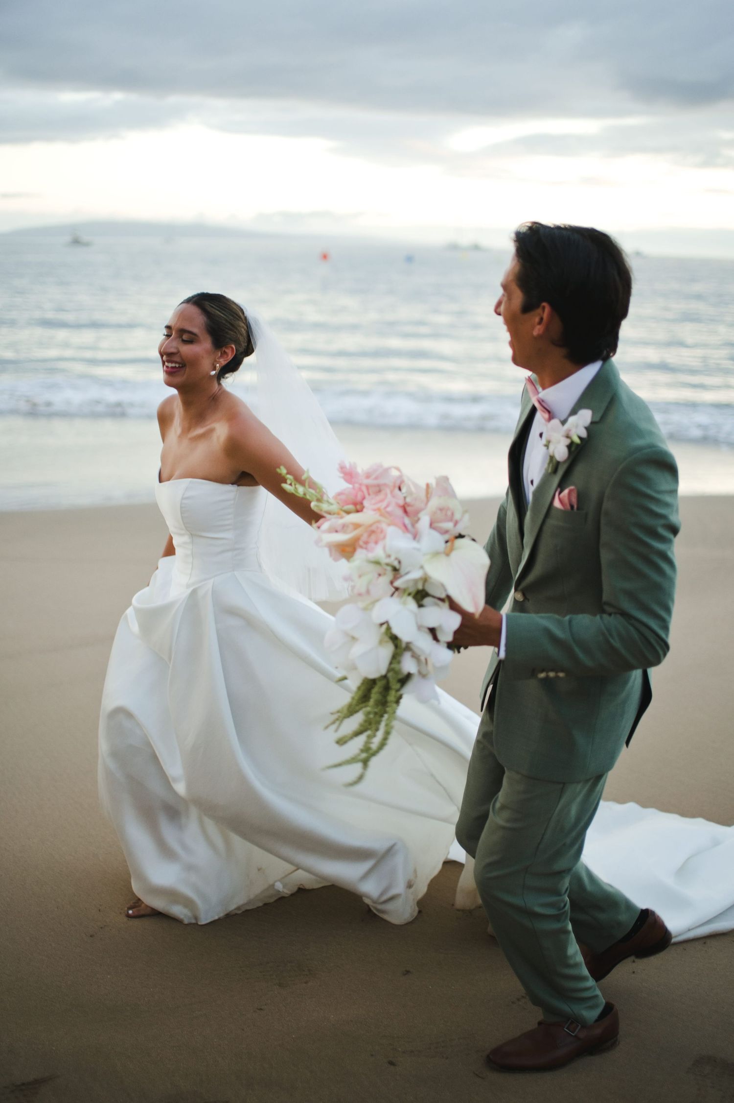 Bride and groom walking hand in hand on a beach, she in a white gown holding pink florals, he in a sage green suit.