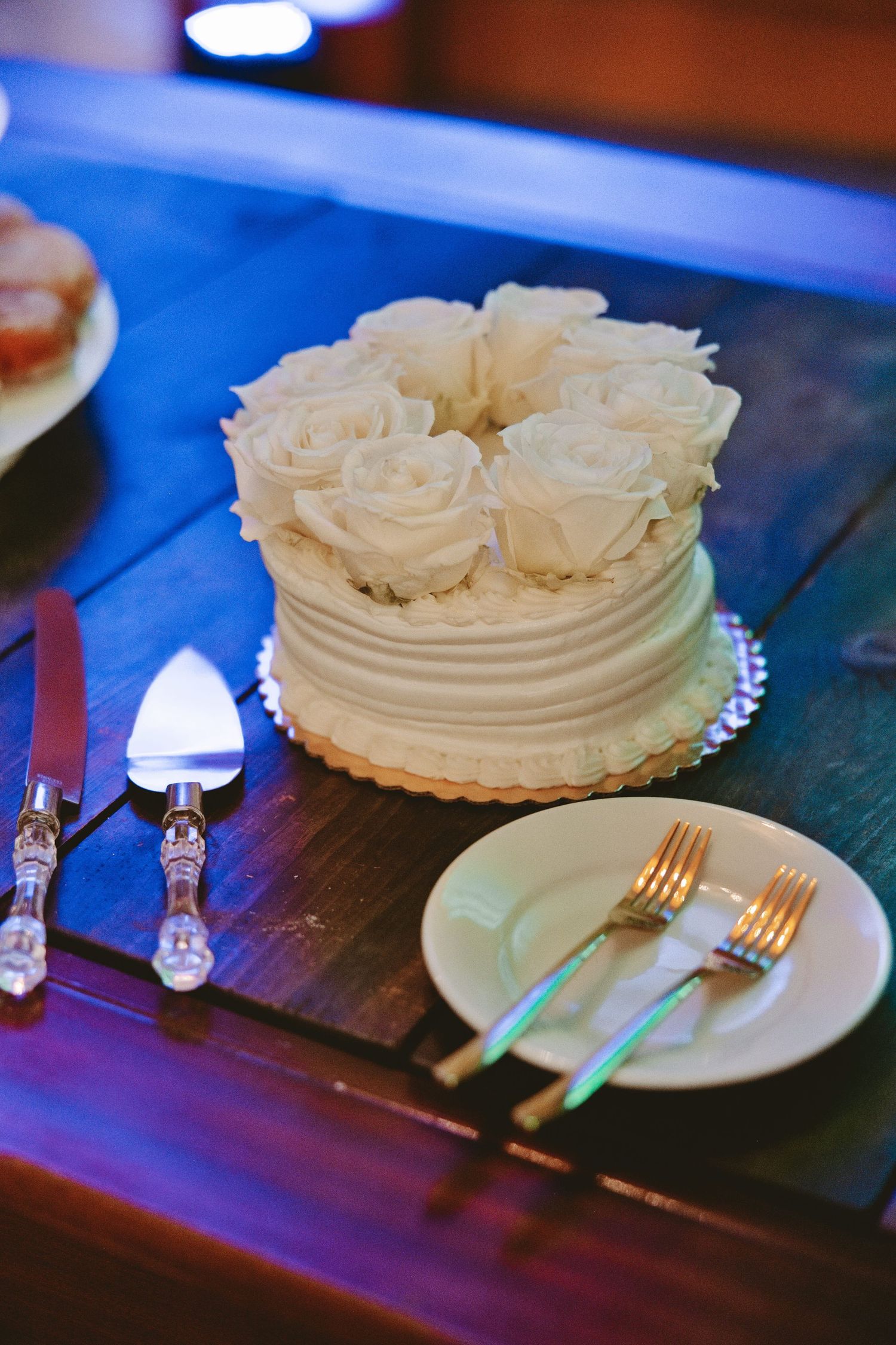 White wedding cake topped with roses on rustic table with cake server and forks.