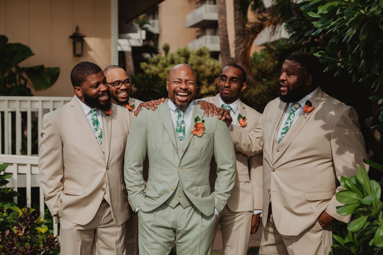 Groom in mint green suit laughing with groomsmen in tan suits and floral ties outdoors.