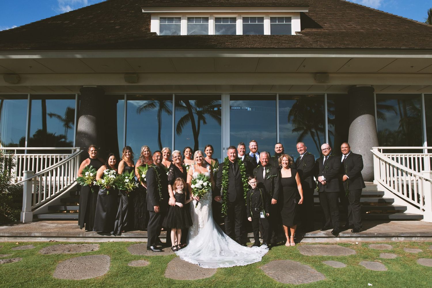 Wedding party in black attire poses on steps of tropical venue with lush greenery and palm trees.