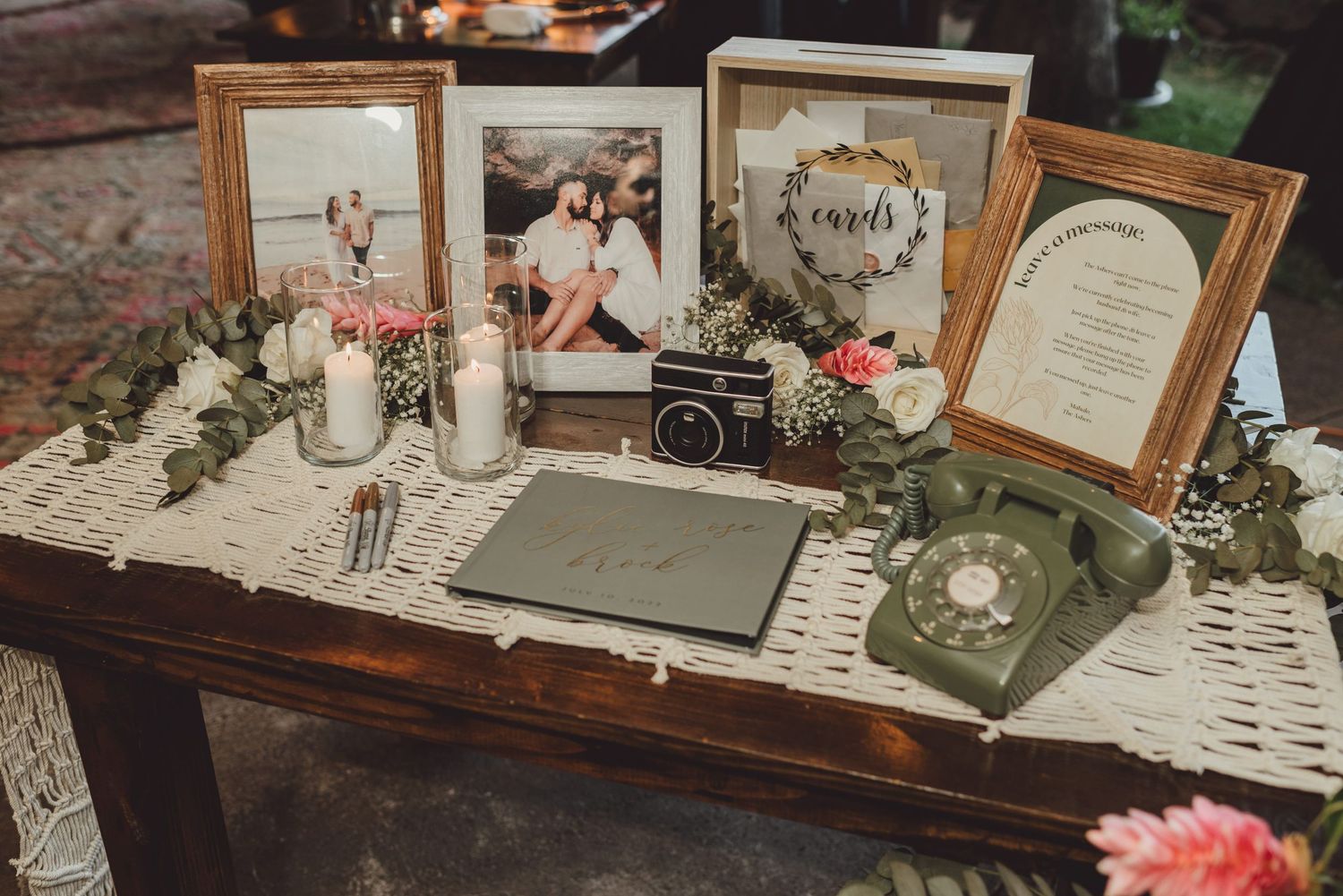 Rustic wedding welcome table with framed photos, candles, greenery, vintage camera, green rotary phone, and guest book.