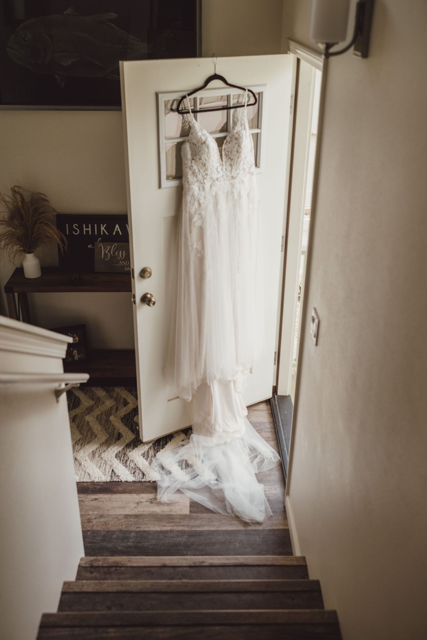 Elegant lace wedding dress hanging on a white door at the top of wooden stairs in a rustic home interior.
