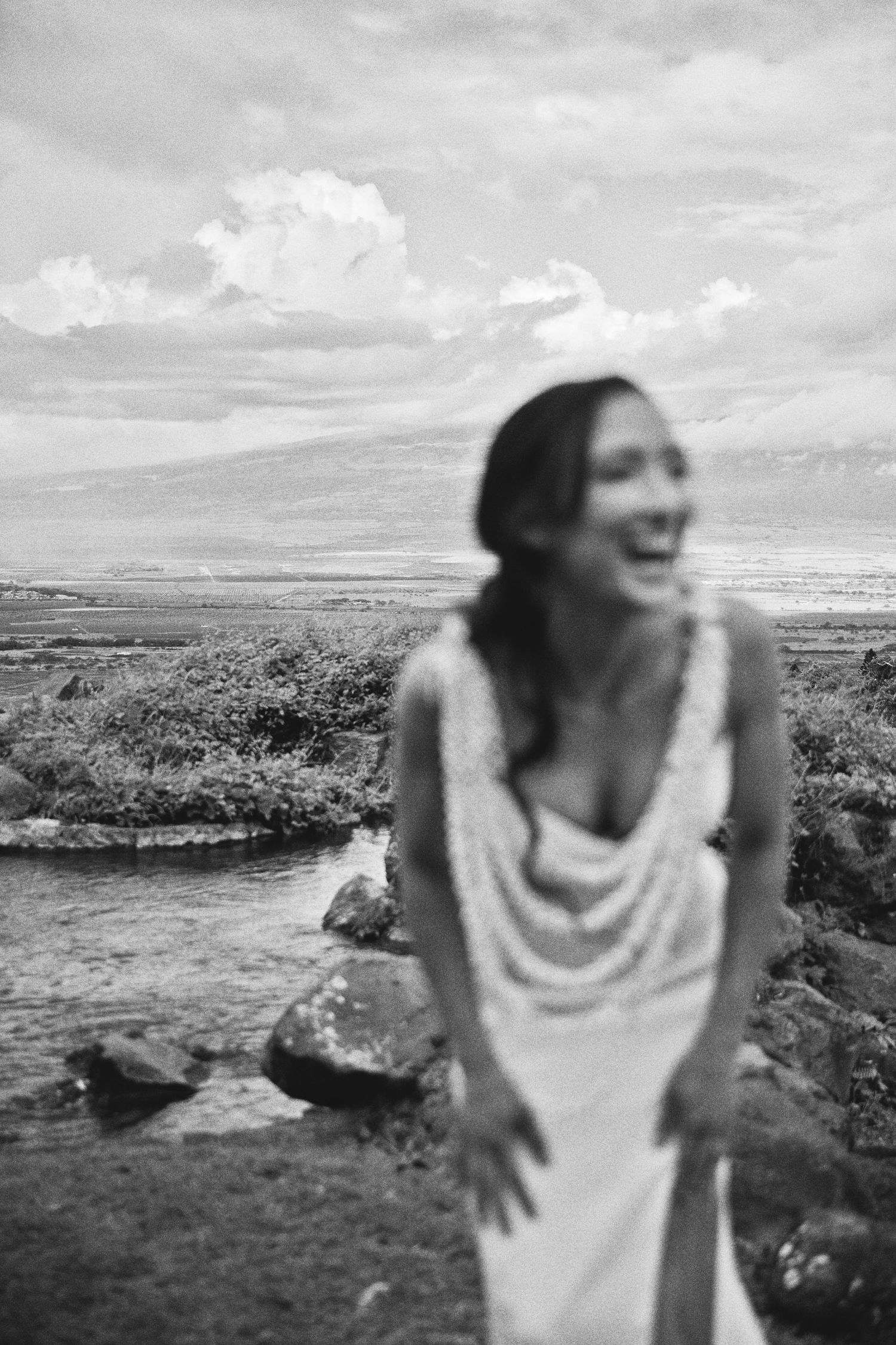 Black and white photo of a laughing bride in a draped gown standing near a rocky stream with mountains in the background.