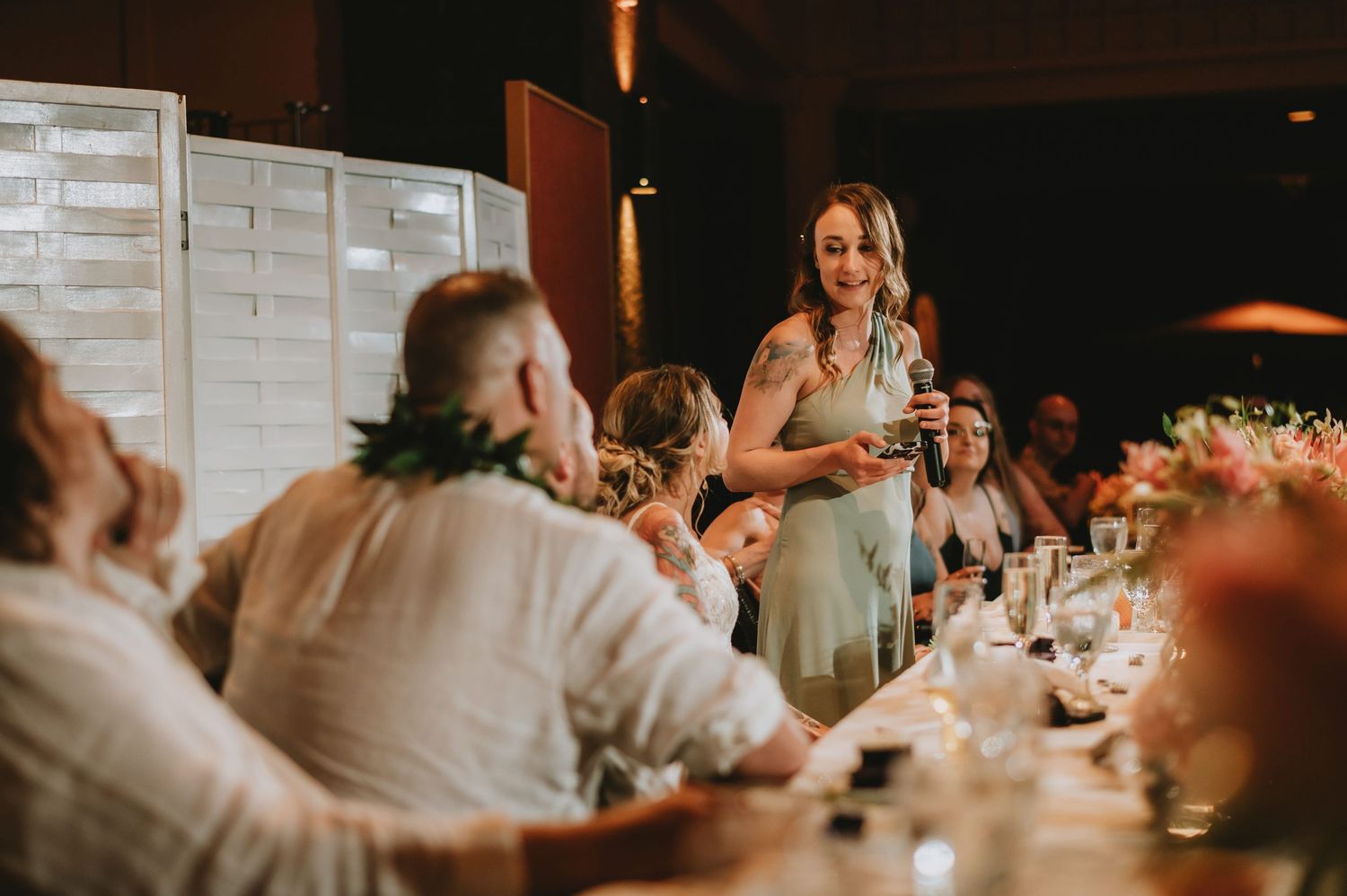 Bridesmaid in sage green dress giving heartfelt toast speech at elegant wedding reception dinner.