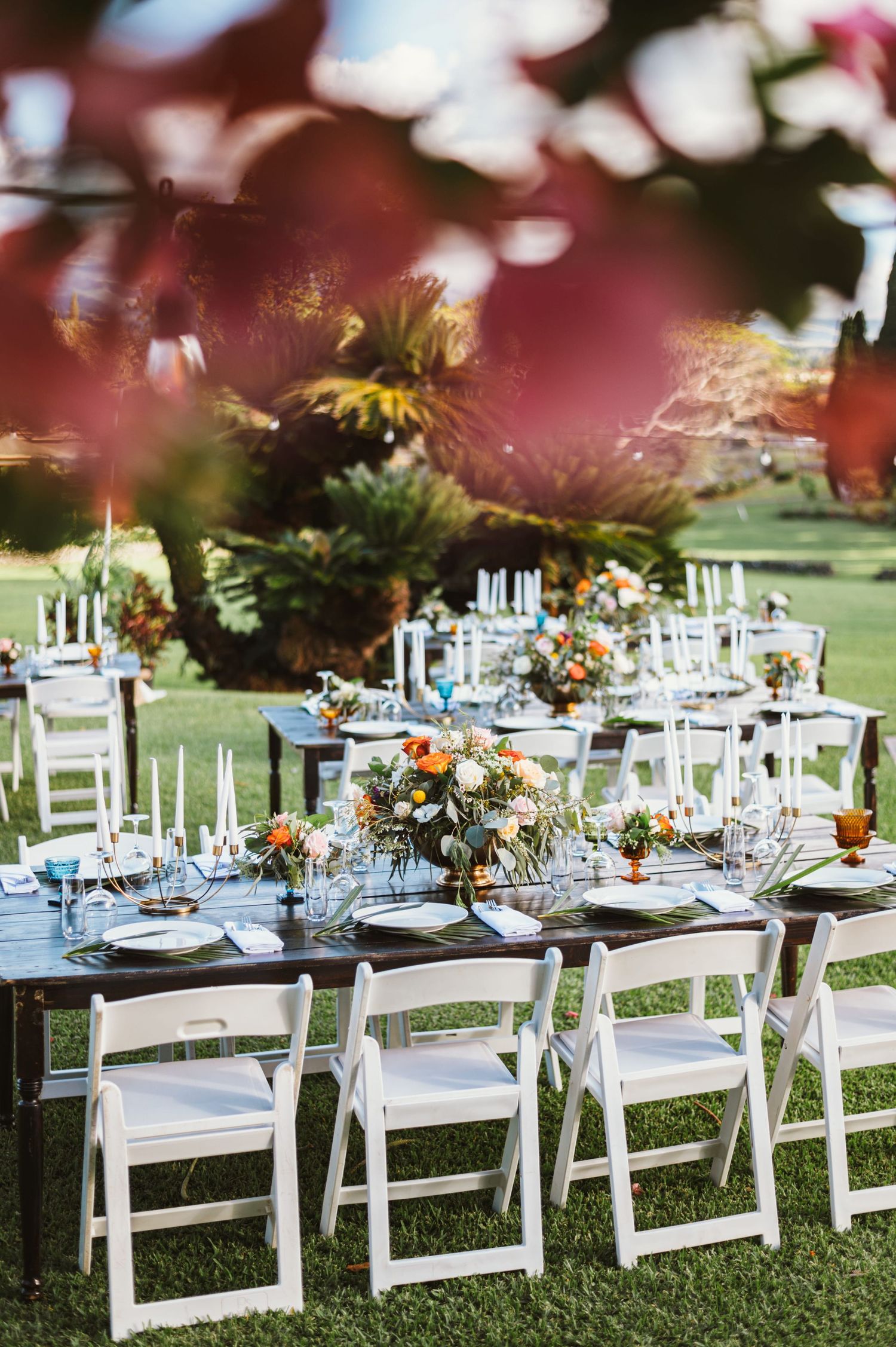 Elegant outdoor wedding reception with white folding chairs, dark wood tables, colorful floral centerpieces, and candles on green lawn.
