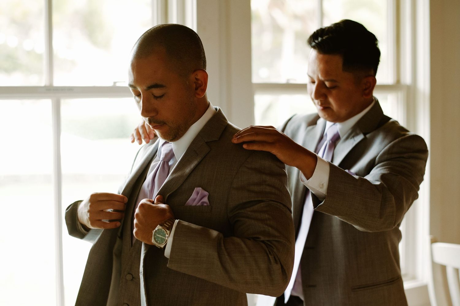 Two men in gray suits adjusting their jackets indoors, preparing for a formal wedding ceremony.