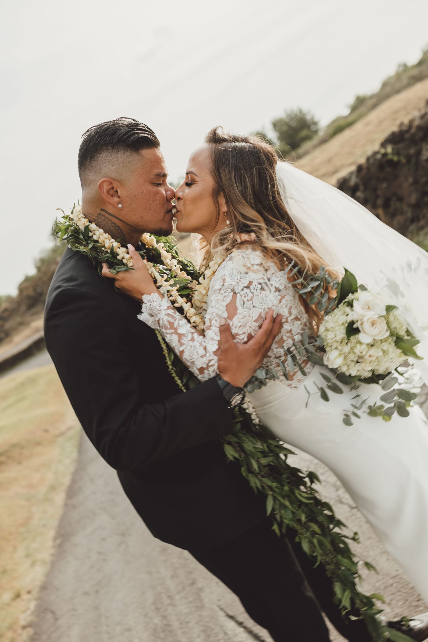 Bride and groom share a romantic kiss outdoors, she holds white floral bouquet, both wearing floral leis on their wedding day.