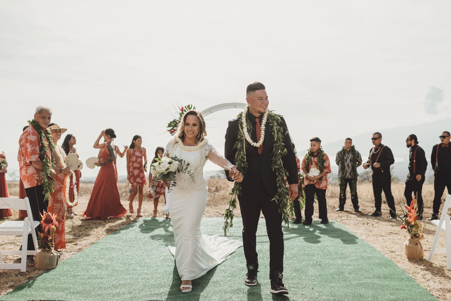 Bride and groom walk down aisle at outdoor wedding ceremony with guests and floral arch backdrop.