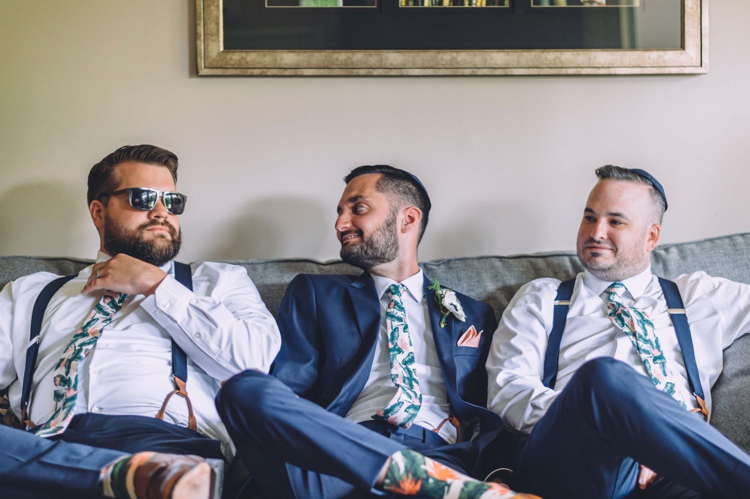 Three groomsmen in matching floral ties relax on a couch during wedding preparations.