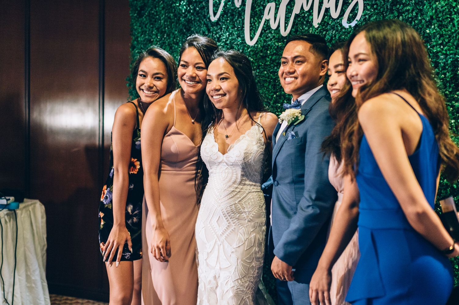 Wedding party poses together against a lush green hedge wall backdrop with decorative signage.