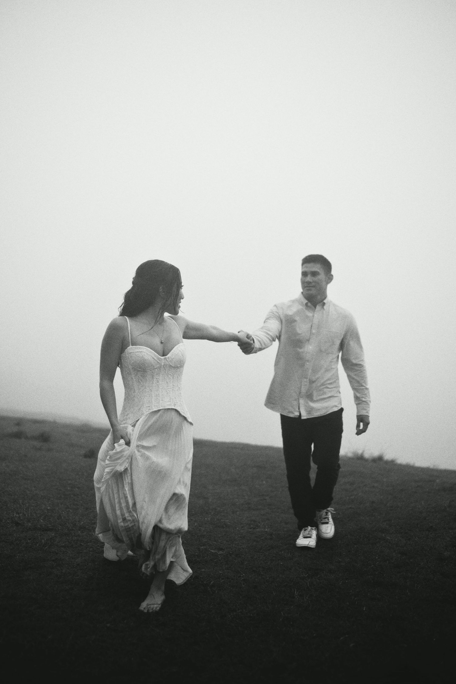 Black and white photo of a couple holding hands and walking through a misty field.