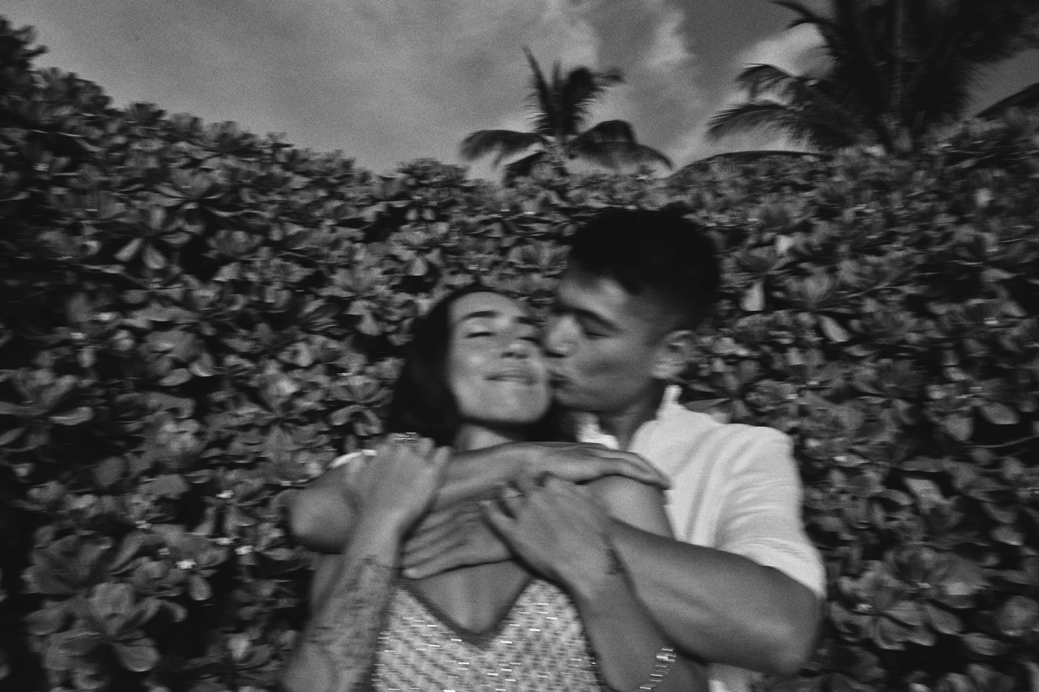 Black and white photo of couple embracing and kissing near tropical foliage.