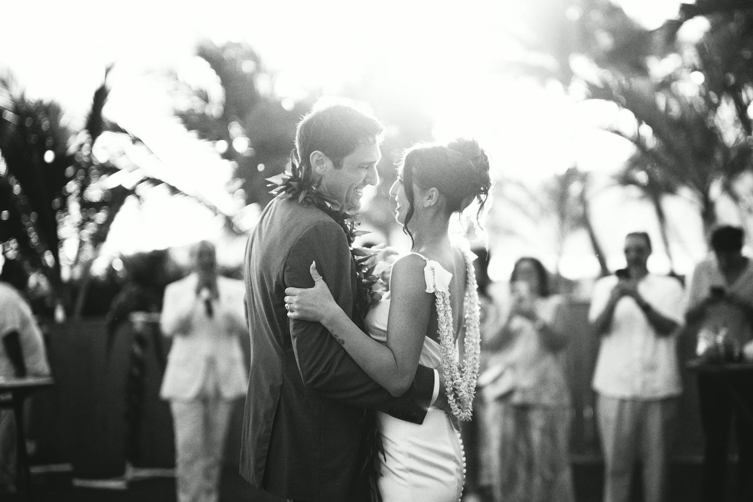 Black and white photo of newlyweds sharing first dance surrounded by guests under tropical palm trees.