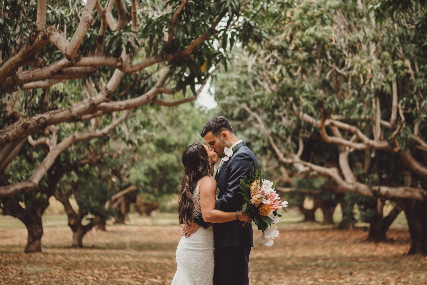 A couple shares a romantic kiss surrounded by lush mango trees, the bride holding a vibrant bouquet.
