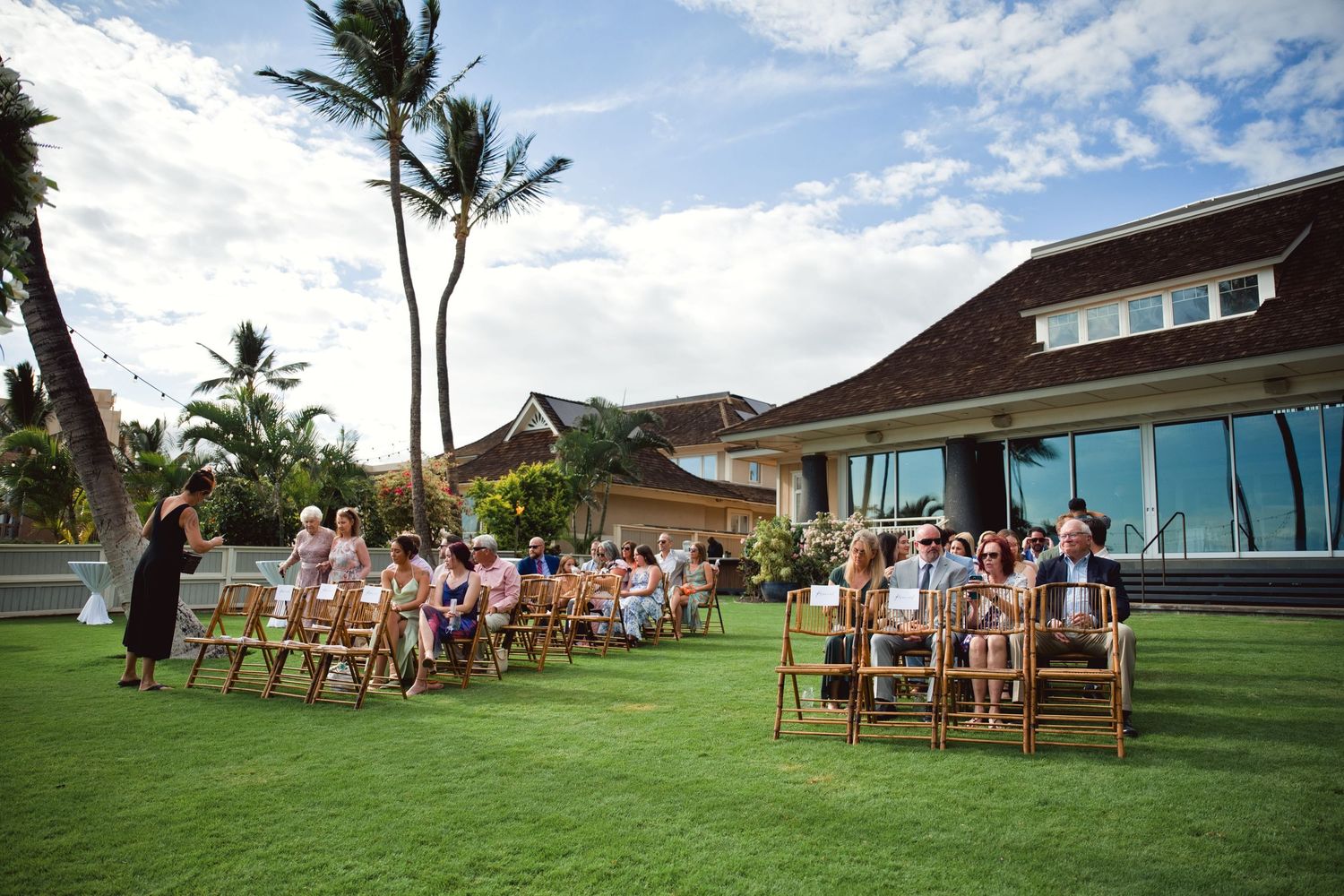 Outdoor wedding ceremony on lush green lawn with wooden chairs, palm trees, and a modern venue in Hawaii.