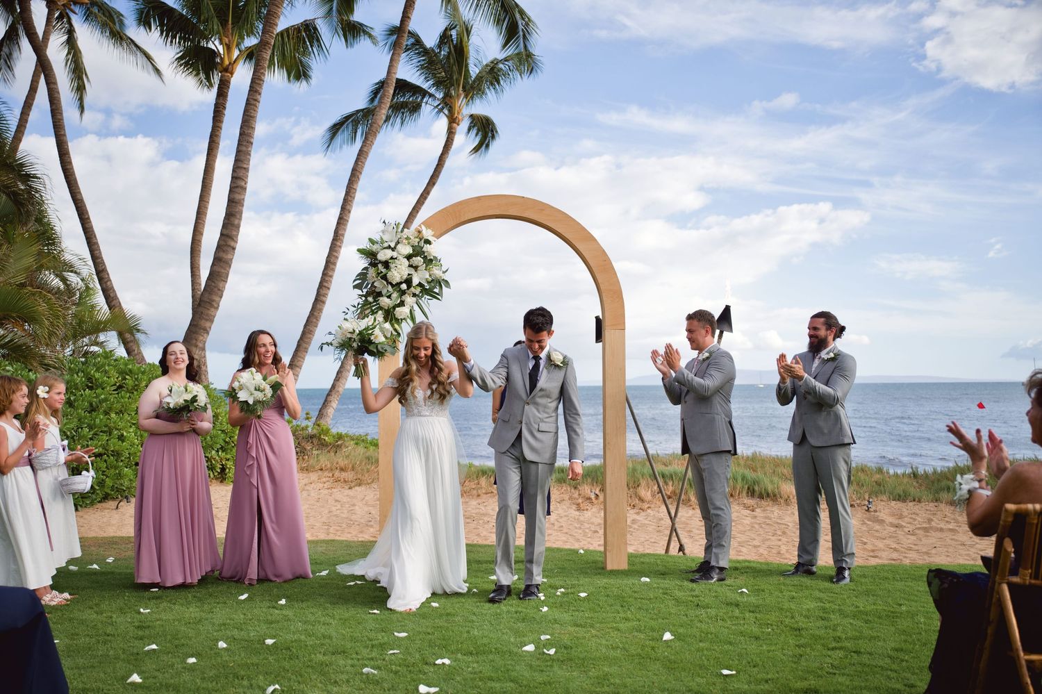 Bride and groom celebrate at outdoor beach wedding ceremony in Maui with palm trees, ocean views, and wooden arch backdrop.