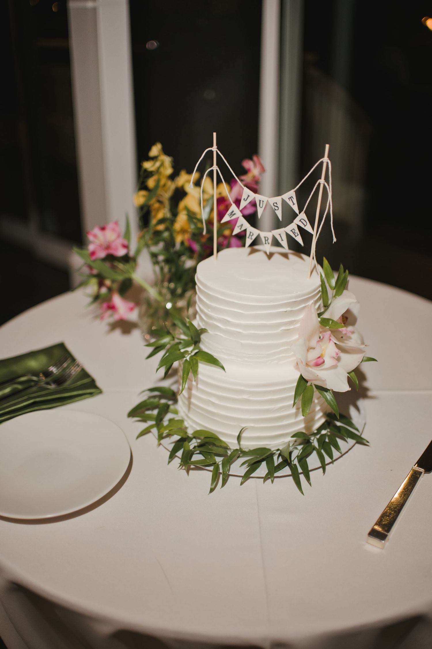 Elegant white tiered wedding cake with greenery wreath, floral topper, and bunting on a white tablecloth.