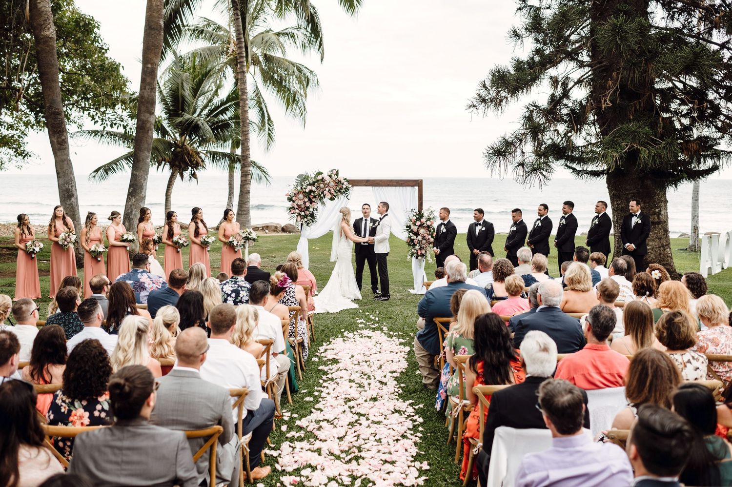 Outdoor beach wedding ceremony with bridesmaids in coral dresses, guests seated on chairs, and couple at floral arch altar.