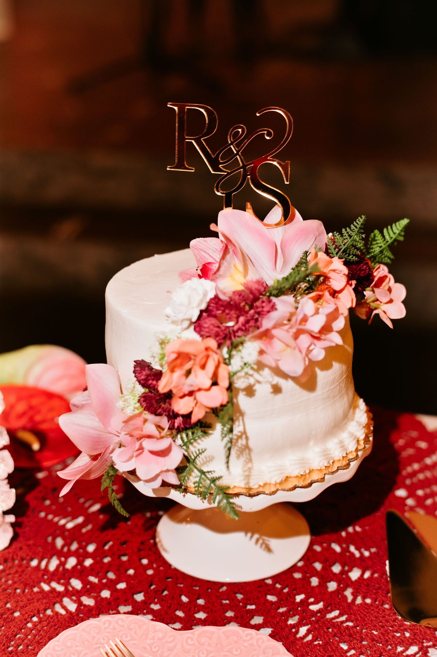 White wedding cake with pink floral decorations and gold R&S monogram topper on red lace tablecloth.