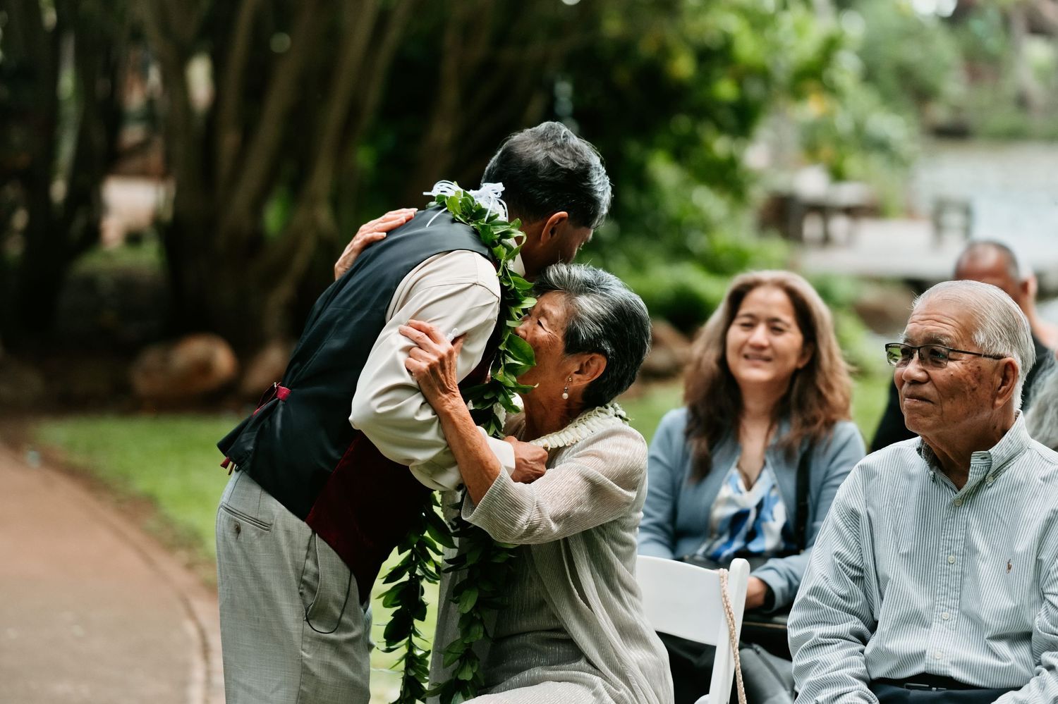 Man wearing lei receives warm hug from elderly woman at outdoor Hawaiian wedding ceremony.