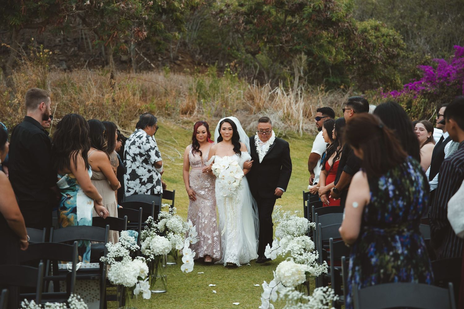 Bride walks down outdoor aisle with escorts past white floral arrangements at a garden wedding ceremony.