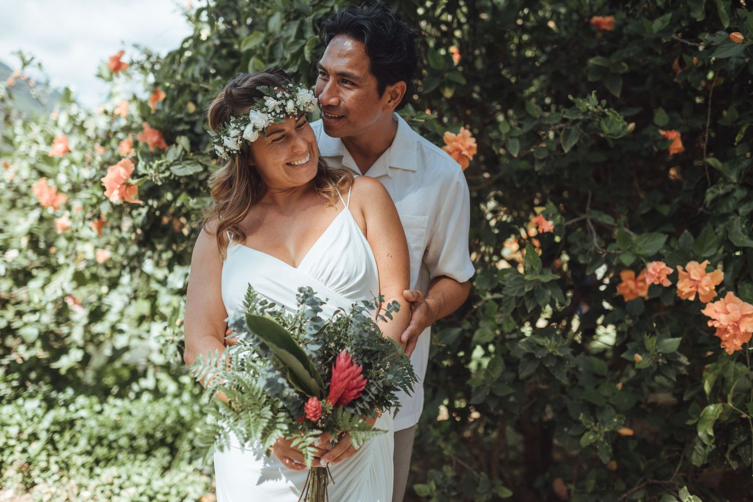 Couple in tropical garden, bride in white dress with floral crown holding bouquet, groom in white shirt smiling.