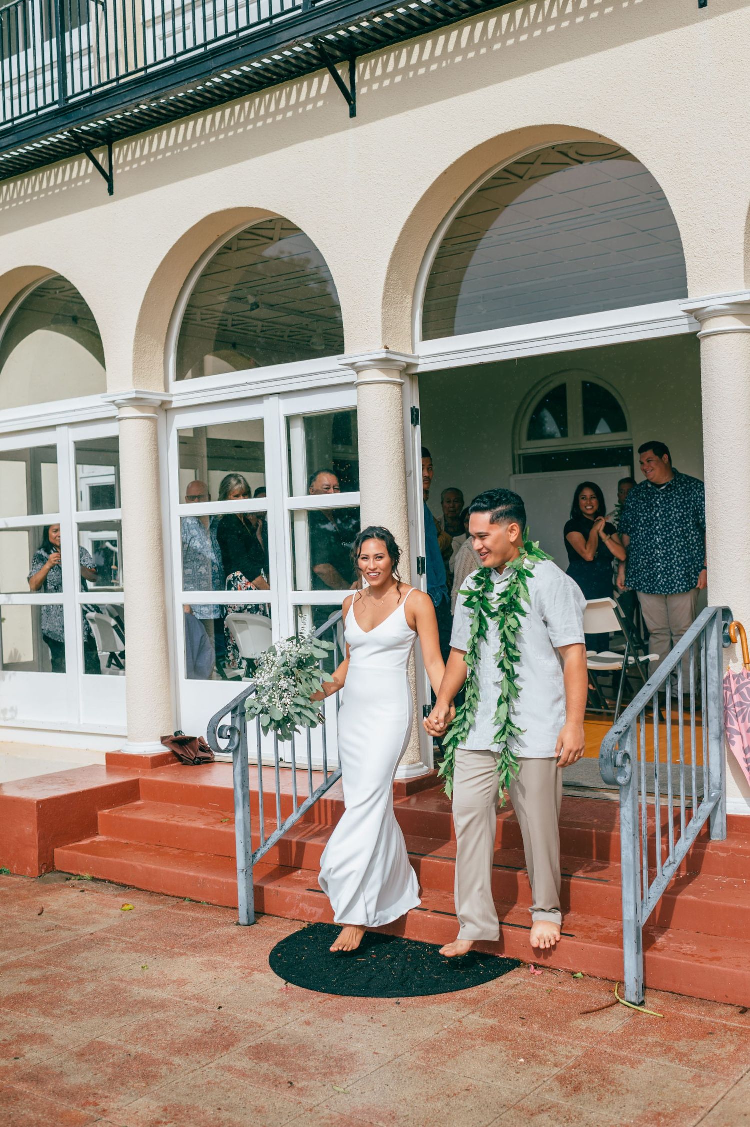 Newlywed couple holding hands walking down steps at Hawaiian wedding venue, groom wearing green lei.