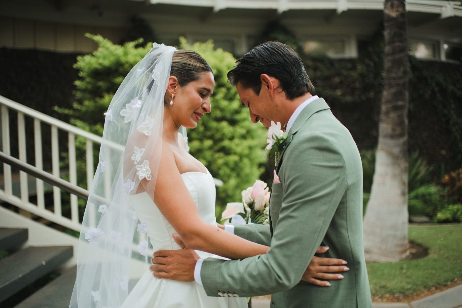 Bride and groom share an intimate moment outdoors, with the bride in a white gown and veil, groom in a sage green suit.