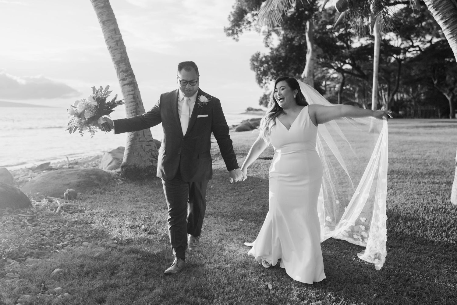 Black and white photo of a joyful bride and groom walking hand in hand outdoors near palm trees on their wedding day.