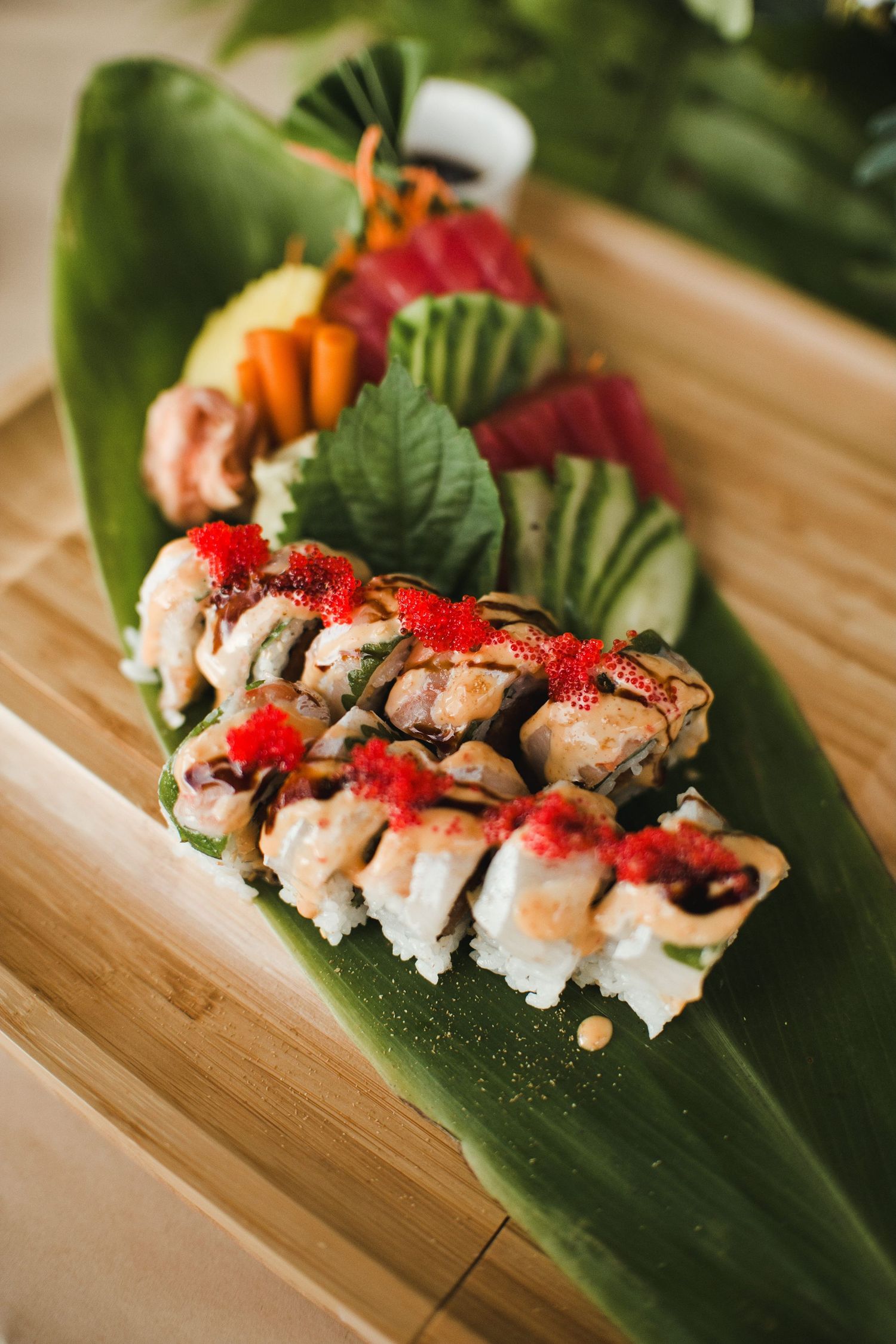 Fresh sushi rolls topped with red tobiko and drizzle sauce, served on a banana leaf with sashimi and garnishes.