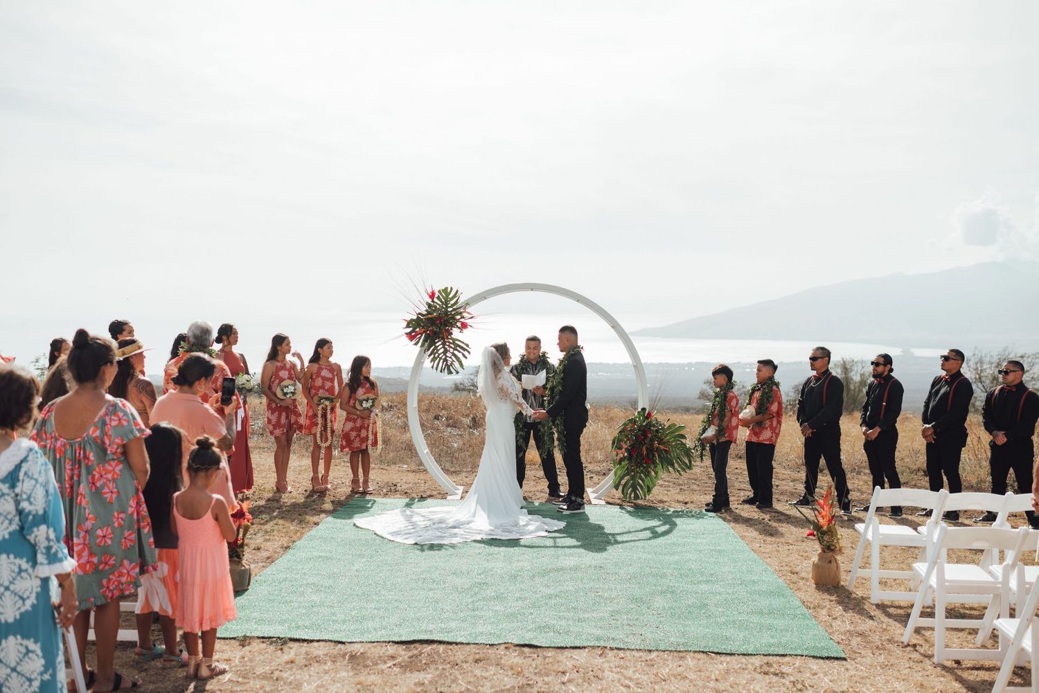 Outdoor beach wedding ceremony with couple at floral arch, guests on both sides, on a sandy shore.