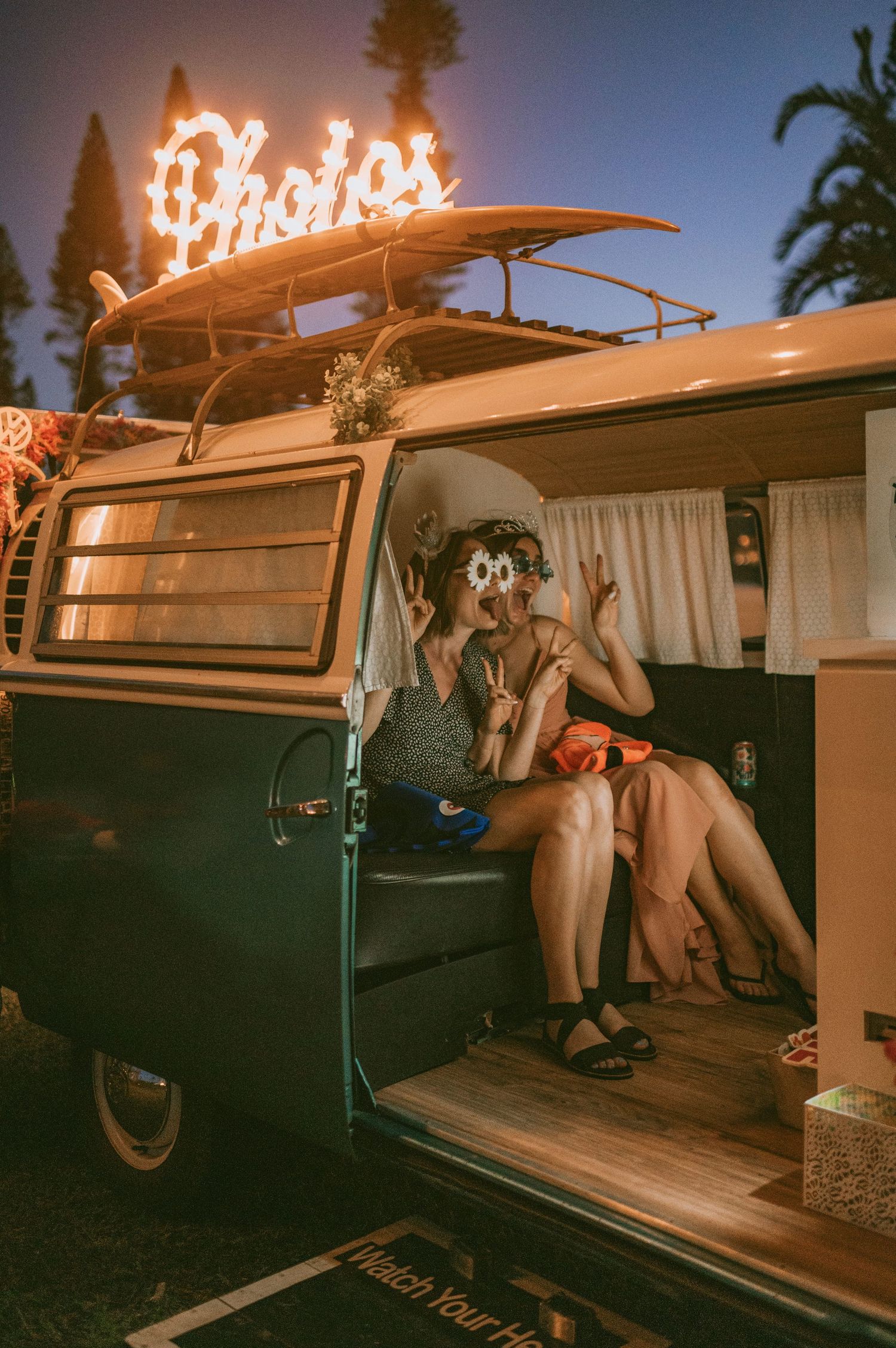 Couple posing in vintage VW bus photo booth at night with neon Photos sign overhead.
