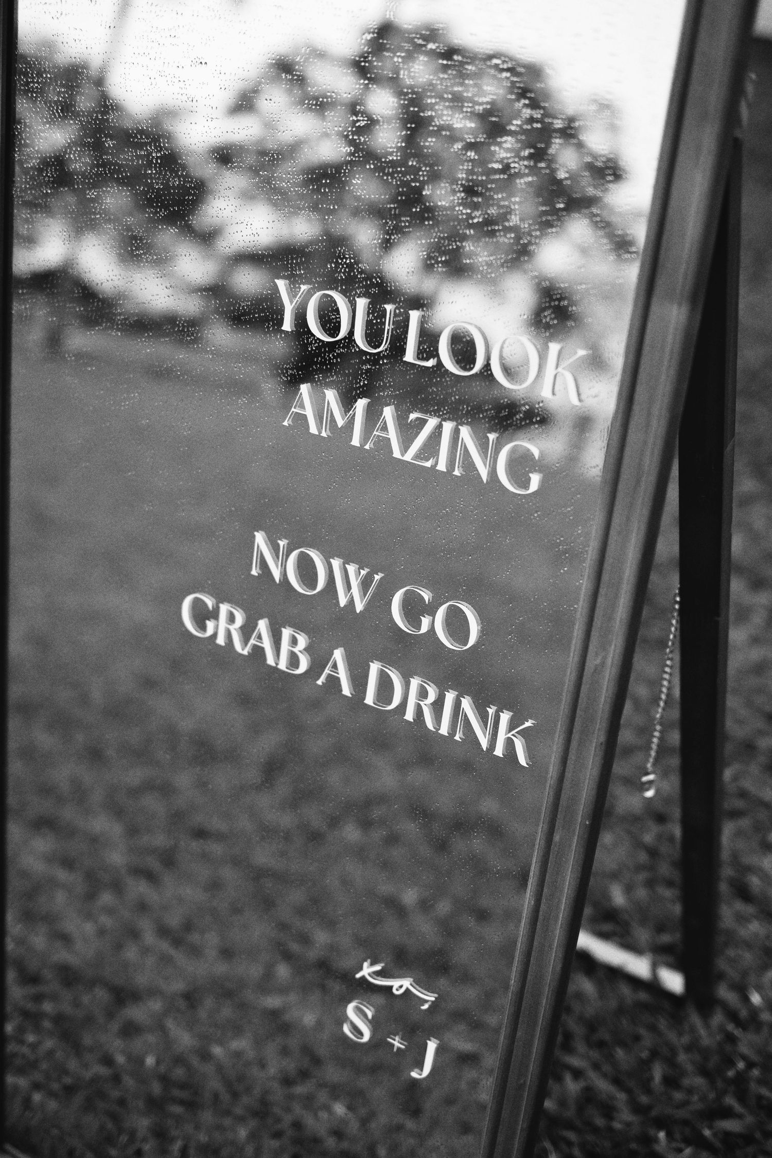 Black and white photo of a decorative mirror sign reading You Look Amazing Now Go Grab A Drink at a wedding.